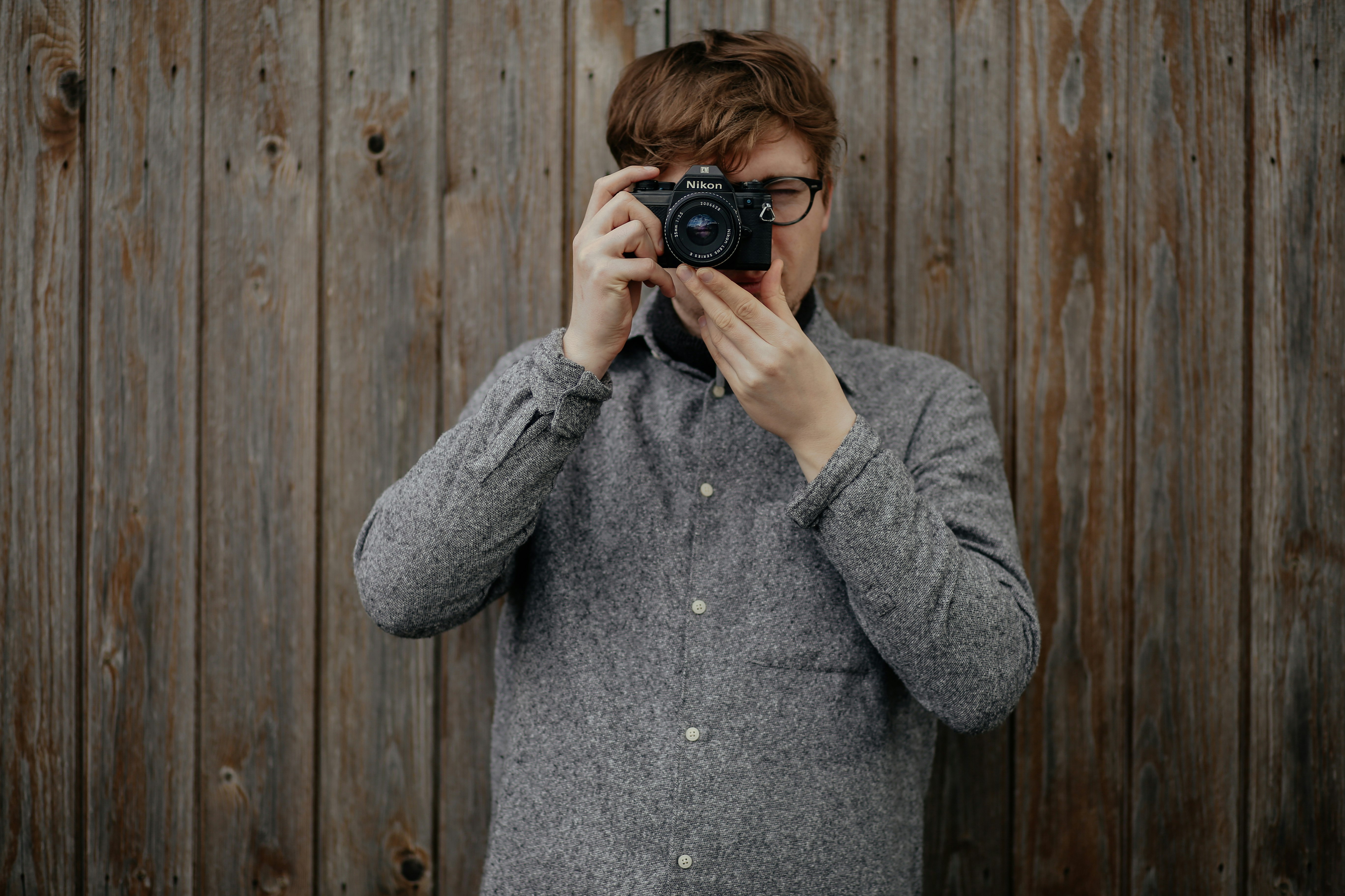 man holding black bridge camera taking photo and standing near brown wooden wall
